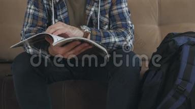 一个男人坐在沙发上看杂志或<strong>手册</strong>，旁边是一个大型<strong>旅游</strong>背包。
